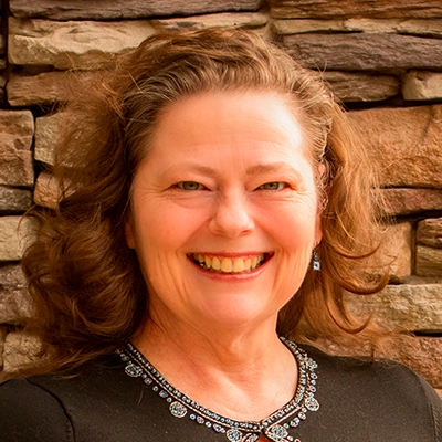 Person with wavy brown hair wearing a black top with decorative beading around the neckline, standing in front of a wall made of stacked stone in shades of brown and tan
