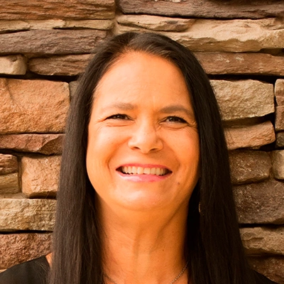 Person with straight dark hair wearing a black top, standing in front of a wall made of stacked stone in shades of brown and tan