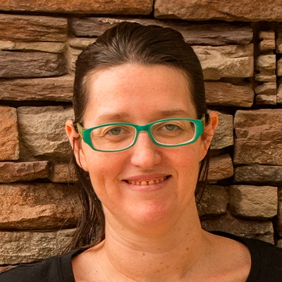 Person with dark brown hair pulled back, wearing a black top and small green earrings, standing in front of a wall made of stacked stone in shades of brown and tan