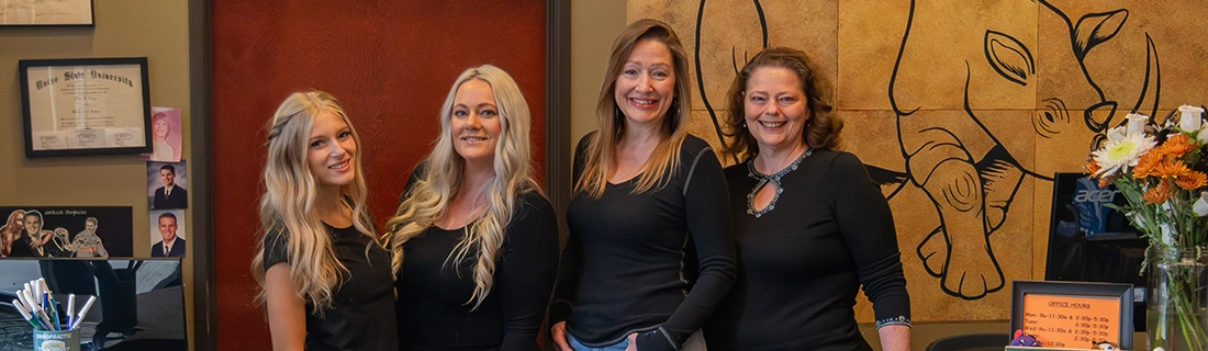 Four people standing indoors in front of a wall with a large rhino artwork. They are wearing black tops and are positioned near a counter with flowers and a framed sign