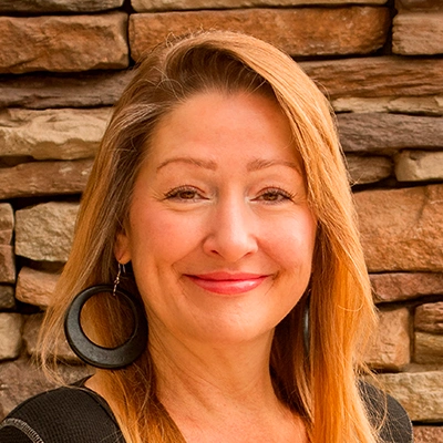 Person with long, straight light brown hair wearing a black top and large circular black earrings, standing in front of a wall made of stacked stone in shades of brown and tan