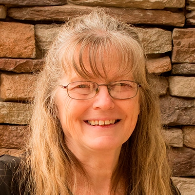 Person with long, wavy light brown hair wearing a black top, standing in front of a wall made of stacked stone in shades of brown and tan