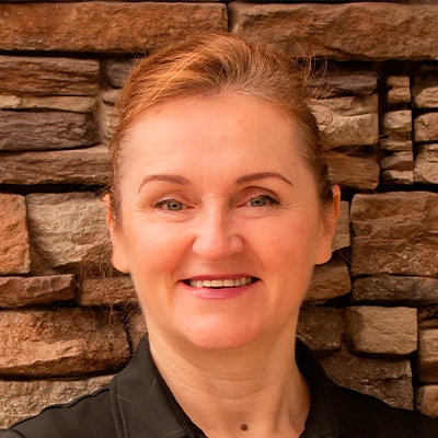 Person wearing a black collared shirt standing in front of a wall made of stacked stone in various shades of brown and tan