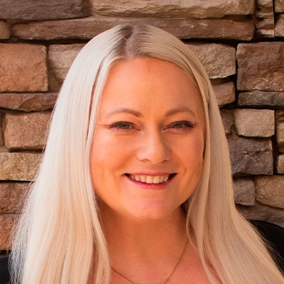 Person with long, straight platinum blonde hair wearing a black top and a delicate gold necklace, standing in front of a wall made of stacked stone in shades of brown and tan
