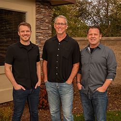Dr. Mick Tiegs & Dr. W. Kevin Davis & Dr. Brendon Smith standing outdoors in front of a stone wall and window, wearing casual button-up shirts and jeans. There are trees and greenery in the background