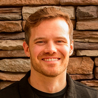 Dr Brendon Smith wearing a black jacket over a dark shirt standing in front of a wall made of stacked stone in shades of brown and tan