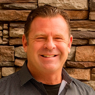 Dr Mick Tiegs wearing a dark patterned button-up shirt standing in front of a wall made of stacked stone with various shades of brown and tan