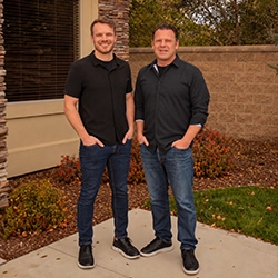 Dr. Mick Tiegs and Dr. Brendon Smith standing outdoors in front of a stone wall and building facade.